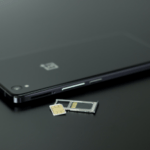 A sleek black smartphone with its SIM card tray ejected, showing a nano-SIM card next to a micro-SIM adapter and a standard SIM card on a dark surface.