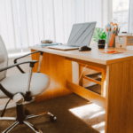 Alt tag: A sunlit home office featuring a wooden desk with an open laptop, a black ergonomic chair, stationery holders, and a small potted plant, with vertical blinds partially closed in the background.