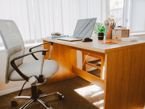 Alt tag: A sunlit home office featuring a wooden desk with an open laptop, a black ergonomic chair, stationery holders, and a small potted plant, with vertical blinds partially closed in the background.