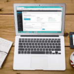 Top-down view of a workspace with an open laptop displaying a webpage, alongside a notepad, a pen, a mobile phone, and a cup of coffee on a wooden desk.