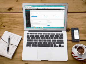 Top-down view of a workspace with an open laptop displaying a webpage, alongside a notepad, a pen, a mobile phone, and a cup of coffee on a wooden desk.