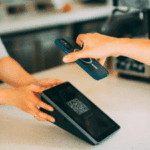 Alt text: A person's hand holding a smartphone above a digital payment terminal with a QR code, indicating a contactless transaction in progress.