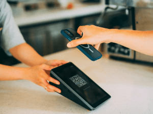 Alt text: A person's hand holding a smartphone above a digital payment terminal with a QR code, indicating a contactless transaction in progress.