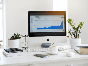 A modern workspace with an iMac displaying a graph on the screen, surrounded by plants, a notebook, a smartphone, and a coffee cup on a white desk.