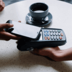 Person making a contactless payment with a smartphone over a card reader with a cup of coffee in the background on a table.