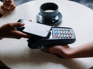 Person making a contactless payment with a smartphone over a card reader with a cup of coffee in the background on a table.