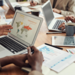 Alt text: A business team analyzes data on a laptop with a world map infographic, surrounded by documents, laptops, and coffee mugs on a conference table.