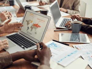 Alt text: A business team analyzes data on a laptop with a world map infographic, surrounded by documents, laptops, and coffee mugs on a conference table.