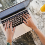 A person typing on a laptop with a marble background accompanied by a glass of orange juice on the right.