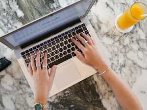 A person typing on a laptop with a marble background accompanied by a glass of orange juice on the right.
