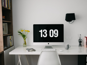 A modern home office setup with a clean white desk featuring an iMac displaying the time 13:09, a black wall-mounted lamp, a vase of yellow flowers, and some office supplies, with bookshelves in the background.