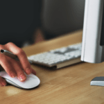 Close-up of a person's hand using a white computer mouse on a wooden desk next to a keyboard and part of a monitor.