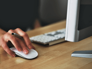 Close-up of a person's hand using a white computer mouse on a wooden desk next to a keyboard and part of a monitor.