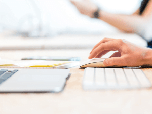 Person working on a computer with documents and a tablet on the desk.