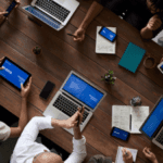 A top-down view of a professional meeting, with multiple people using laptops and digital tablets around a wooden table, with notes and a small plant visible.