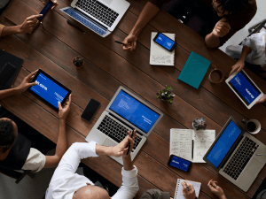 A top-down view of a professional meeting, with multiple people using laptops and digital tablets around a wooden table, with notes and a small plant visible.