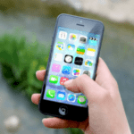 A hand holding a smartphone with colorful app icons on the screen, blurred greenery in the background.