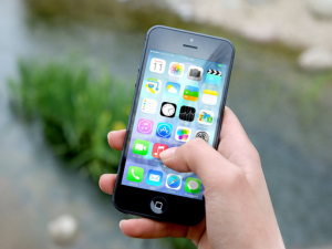 A hand holding a smartphone with colorful app icons on the screen, blurred greenery in the background.