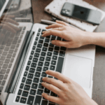 Close-up of a person's hands typing on a laptop keyboard with a smartphone and notepad alongside on a wooden table.