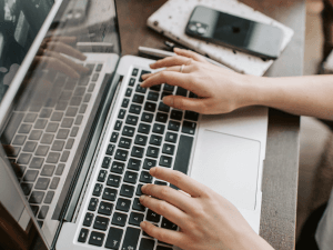 Close-up of a person's hands typing on a laptop keyboard with a smartphone and notepad alongside on a wooden table.