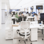 Alt text: Modern office space featuring rows of white desks with computers, ergonomic chairs, and individuals working, accented with green plants and natural light.