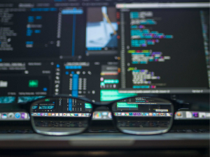 Eyeglasses in the foreground with clear focus on their lenses, showing reflected colorful coding software screens blurred in the background.