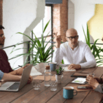 Four professionals engaged in a discussion around a conference table with laptops, notepads, and drinks in a modern office setting.