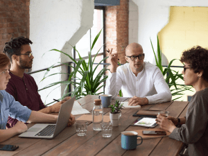 Four professionals engaged in a discussion around a conference table with laptops, notepads, and drinks in a modern office setting.