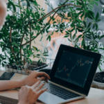 Person analyzing financial charts on a laptop in a cozy workspace surrounded by indoor plants.