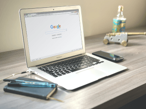 Modern workspace with an open laptop showing Google's homepage, accompanied by a smartphone, notepads, pens, and a decorative glass bottle on a wooden desk.