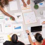 Top-down view of a business team collaborating around a table covered with documents, charts, and a laptop, indicating a strategic planning session.