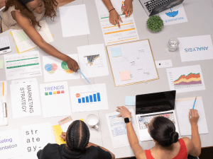 Top-down view of a business team collaborating around a table covered with documents, charts, and a laptop, indicating a strategic planning session.