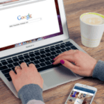 Person using a laptop with Google search homepage on the screen, a smartphone, and a cup of coffee on a wooden desk.