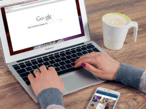 Person using a laptop with Google search homepage on the screen, a smartphone, and a cup of coffee on a wooden desk.