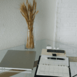 A modern home office setup with a closed laptop, a clear clipboard with papers, glasses, smartphone, pen, and a vase of dried wheat on a glass table against a white brick wall.