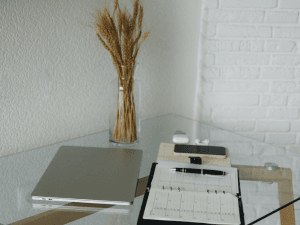 A modern home office setup with a closed laptop, a clear clipboard with papers, glasses, smartphone, pen, and a vase of dried wheat on a glass table against a white brick wall.