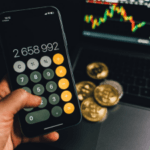 Hand holding a smartphone with a calculator app displayed, showing a large number, with a blurry background featuring a laptop screen with stock market graphs and physical coins.