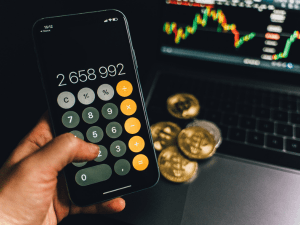 Hand holding a smartphone with a calculator app displayed, showing a large number, with a blurry background featuring a laptop screen with stock market graphs and physical coins.