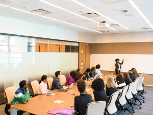 Alt text: A diverse group of professionals attending a presentation by a speaker in a well-lit conference room with large windows.