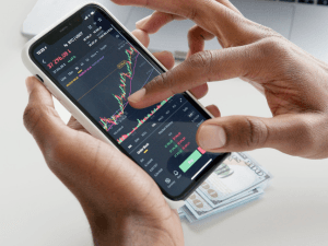 A person's hands holding a smartphone displaying stock market charts, with US dollar bills lying underneath on a table.