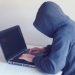 Person in a gray hoodie typing on a laptop with code on the screen, suggesting a programmer or hacker at work, in a room with a white backdrop.