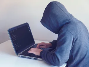 Person in a gray hoodie typing on a laptop with code on the screen, suggesting a programmer or hacker at work, in a room with a white backdrop.
