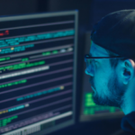 Man wearing glasses and cap focused on code on computer monitors in dimly lit room, reflecting cyber work environment.