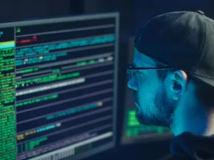 Man wearing glasses and cap focused on code on computer monitors in dimly lit room, reflecting cyber work environment.