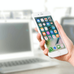 Close-up of a hand holding a smartphone with app icons on the screen, laptop in the blurred background.