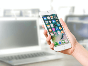 Close-up of a hand holding a smartphone with app icons on the screen, laptop in the blurred background.