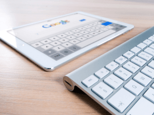 A tablet with a Google search page displayed lies on a wooden desk next to a white Bluetooth keyboard, emphasizing modern wireless technology and internet access.