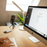 A person working at an organized desk with an iMac displaying a Google search page, a smartphone lying nearby, and a podcasting microphone in the background.