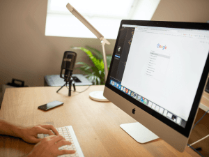 A person working at an organized desk with an iMac displaying a Google search page, a smartphone lying nearby, and a podcasting microphone in the background.