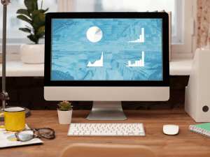 A home office setup featuring a desktop computer with graphs and pie chart on the screen, surrounded by a plant, glasses, and a mug on a wooden desk with white background.